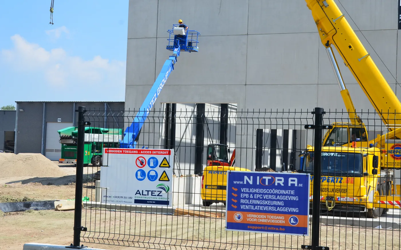 Nitra-werfbord en veiligheidssignalisatie aan de omheining van een werf, met hoogtewerker en kraan